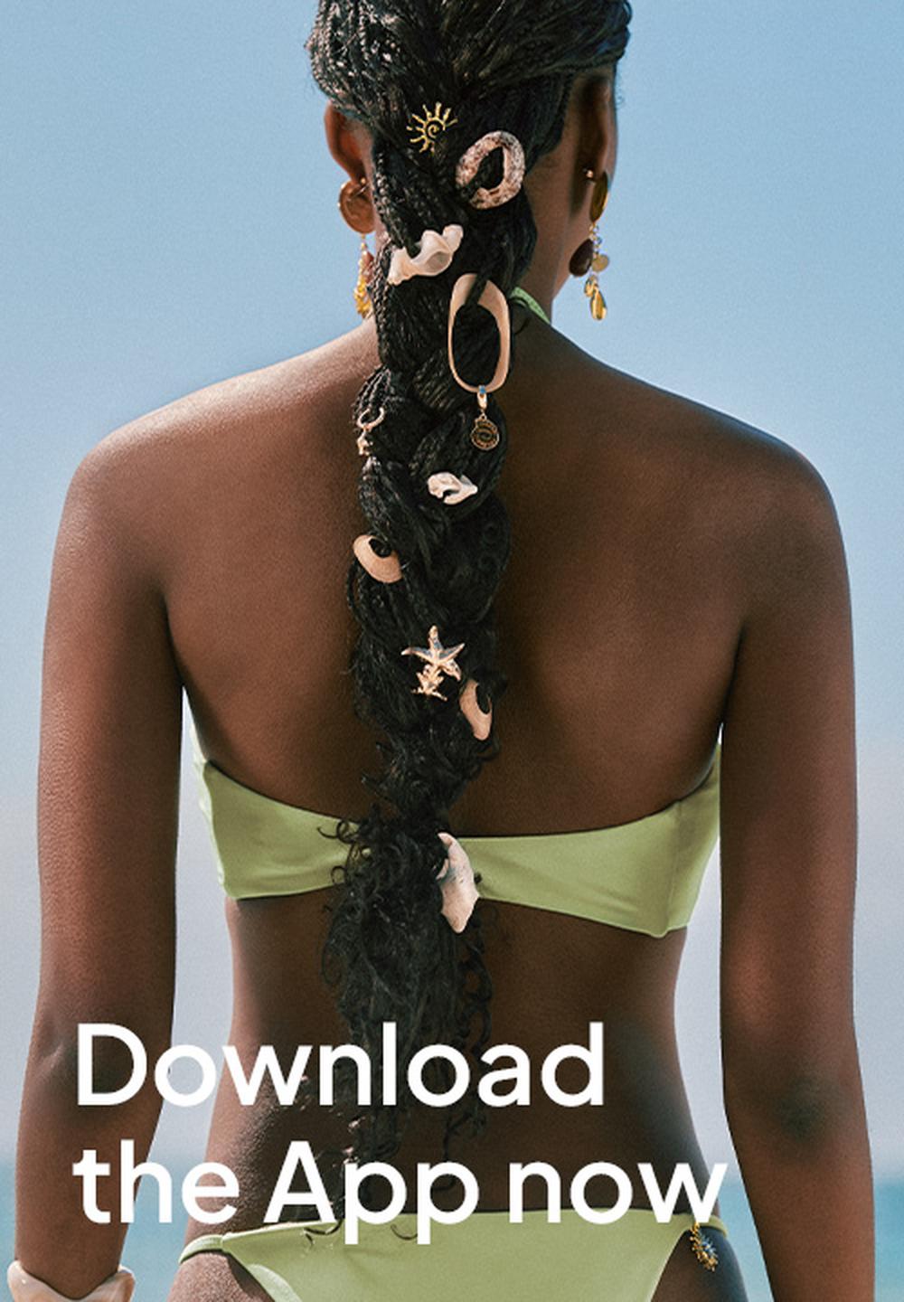 Promotional Superbalist banner showing the back of a woman in a light green bikini against a blue background, with decorative accessories in her braided hair and the overlaid text “Download the App now” in large white letters.