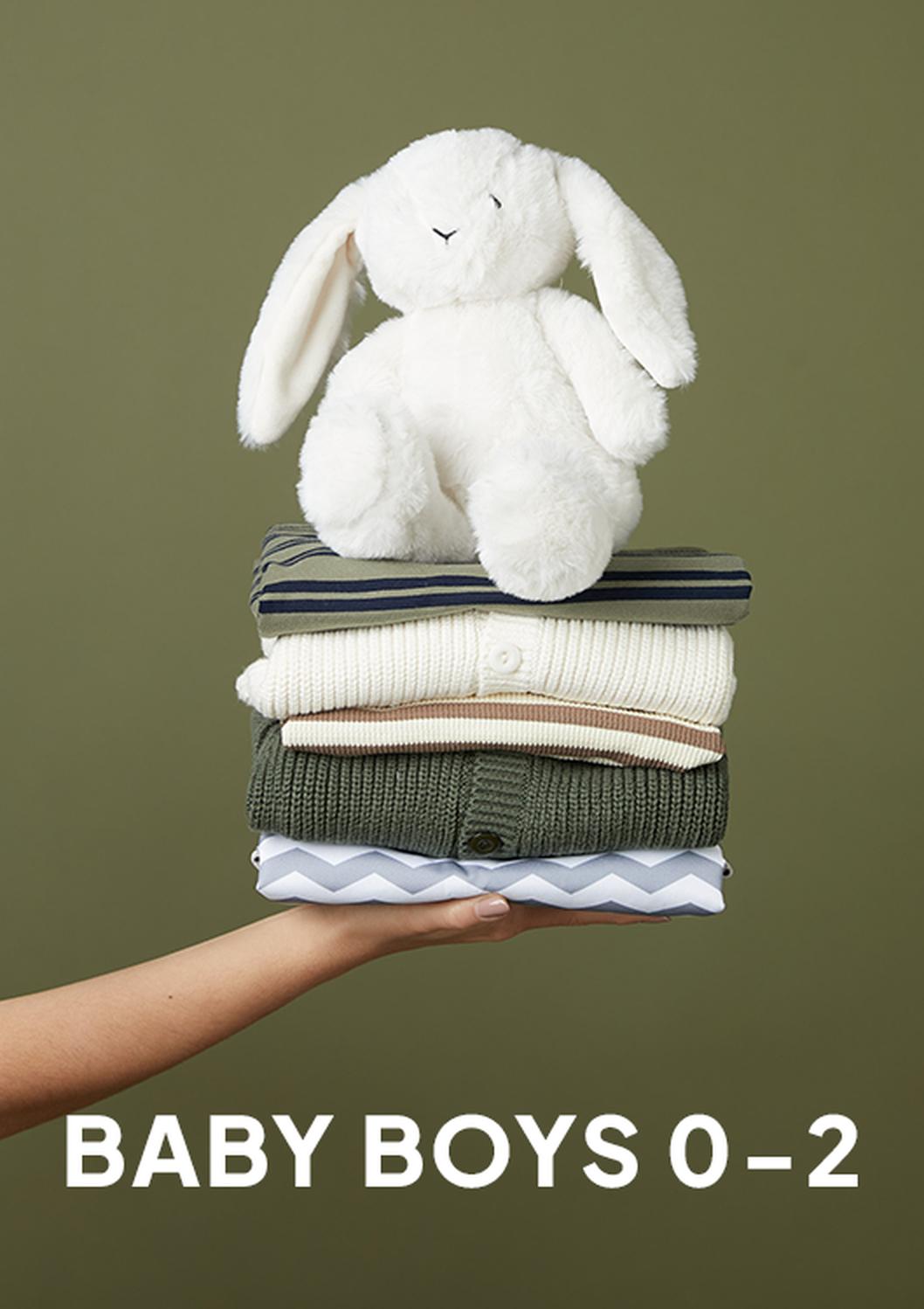 A hand holds a stack of folded baby clothes topped with a white plush bunny against an olive background, with the text “BABY BOYS 0–2” at the bottom.