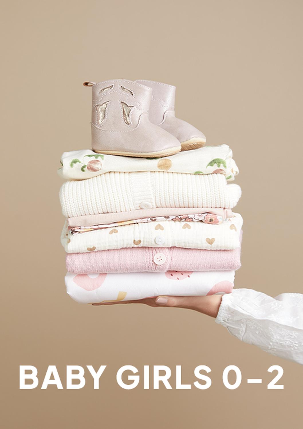 A hand in a white sleeve holds a stack of folded baby girl clothes topped with soft pink baby booties, against a beige background, with the text “BABY GIRLS 0–2” below.