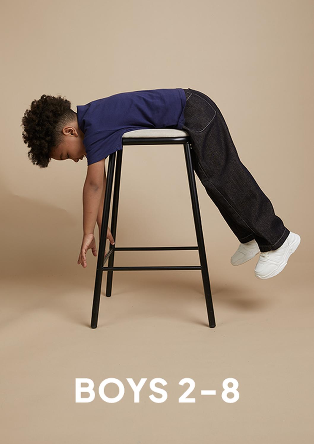 A young boy in a navy shirt, dark jeans, and white sneakers lies draped face-down over a black stool against a tan studio backdrop, with “BOYS 2–8” text below.