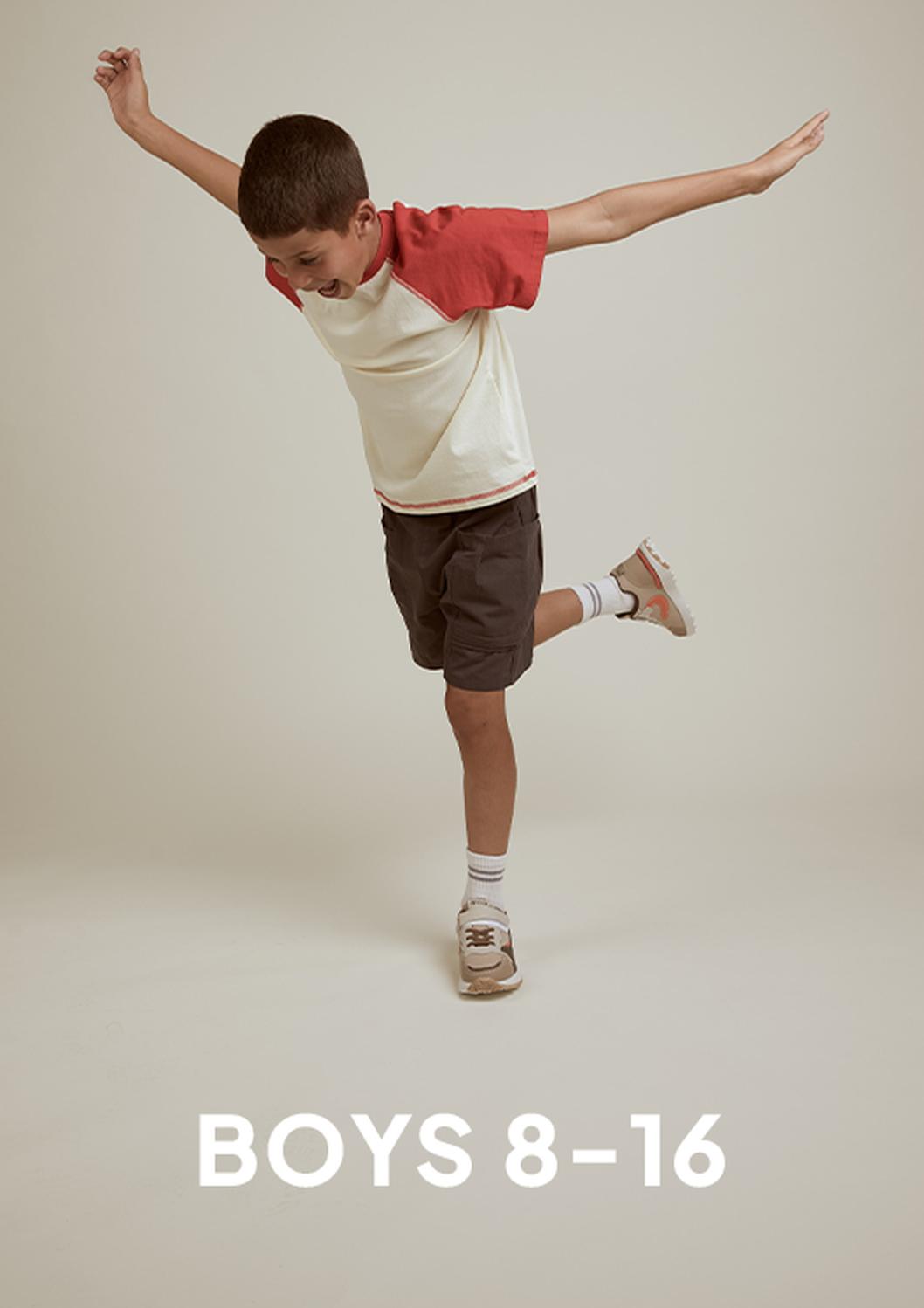 A boy in a cream and red T-shirt, dark shorts, socks, and sneakers balances on one leg with arms outstretched against a beige studio backdrop, with the text “BOYS 8–16” below.