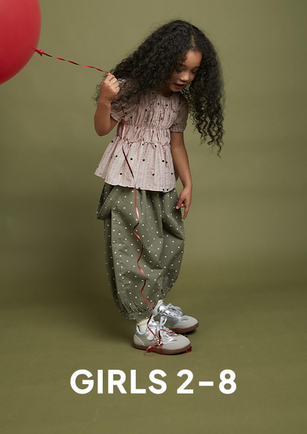 A young girl with long curly hair stands against an olive-green studio backdrop holding a red balloon, wearing a pink top, loose olive polka-dot pants, and silver sneakers, with the text “GIRLS 2–8” below.