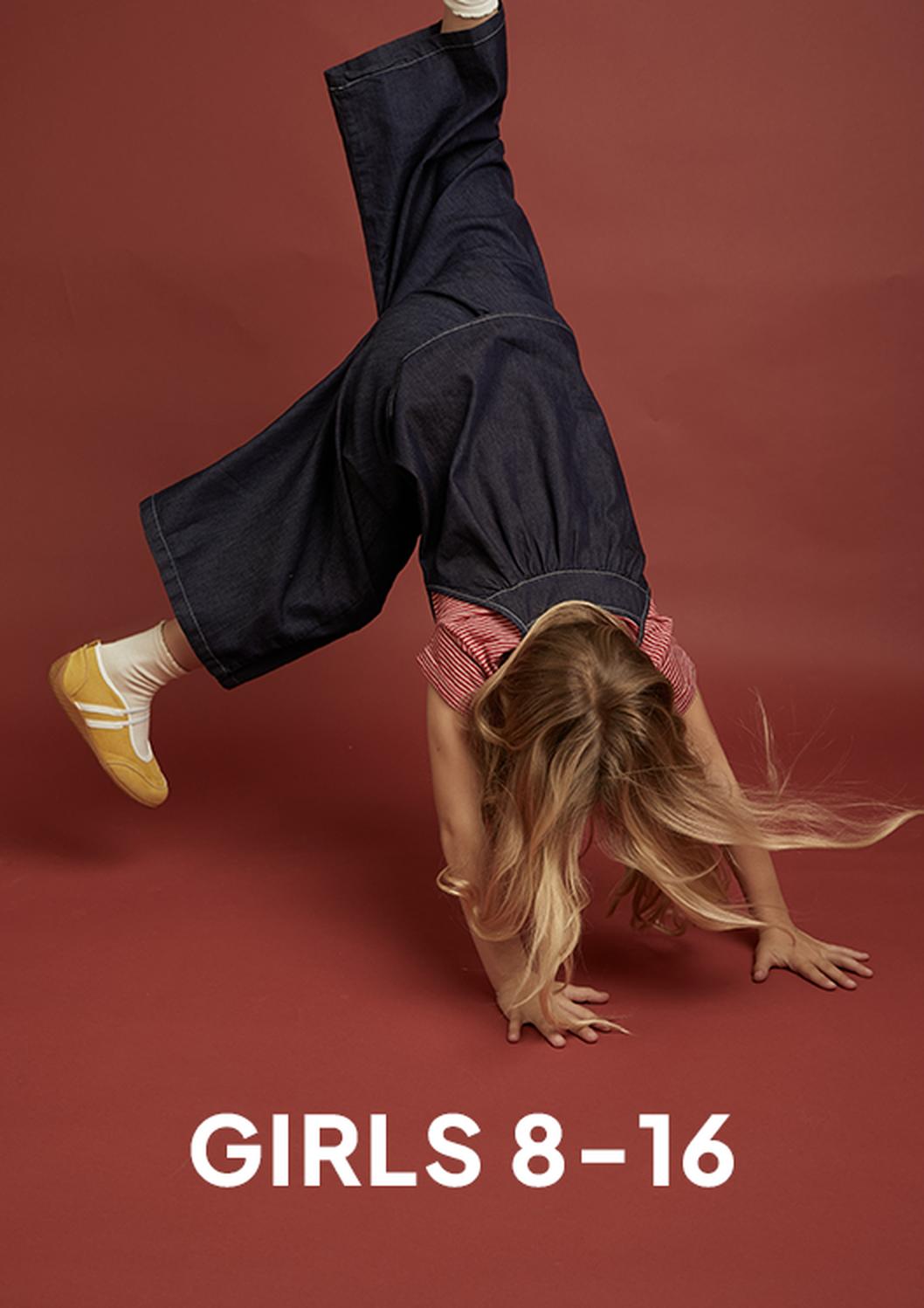 A girl in a red striped top, loose dark jeans, and yellow sneakers does a handstand against a reddish-brown studio backdrop, with the text “GIRLS 8–16” below.