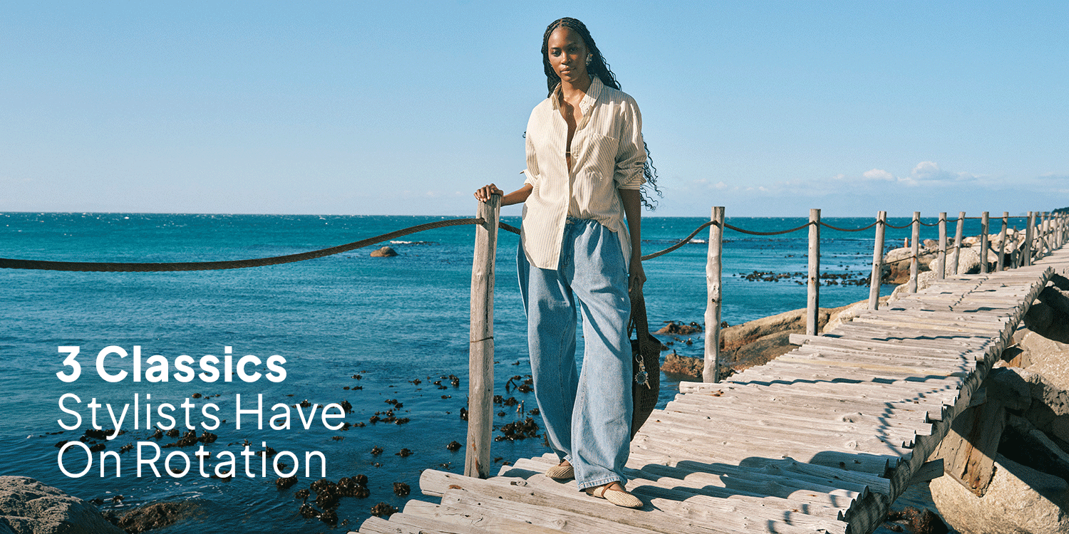 Fashion campaign banner of a woman in a cream shirt and wide-leg blue trousers standing on a wooden seaside bridge, with the text “3 Classics Stylists Have On Rotation.”