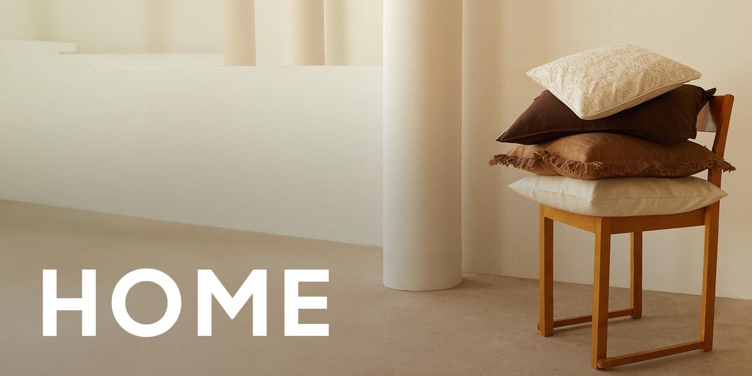 Minimalist beige home interior with stacked cushions on a wooden stool and the word “HOME” overlaid in large white text, representing Black Friday home and living deals.