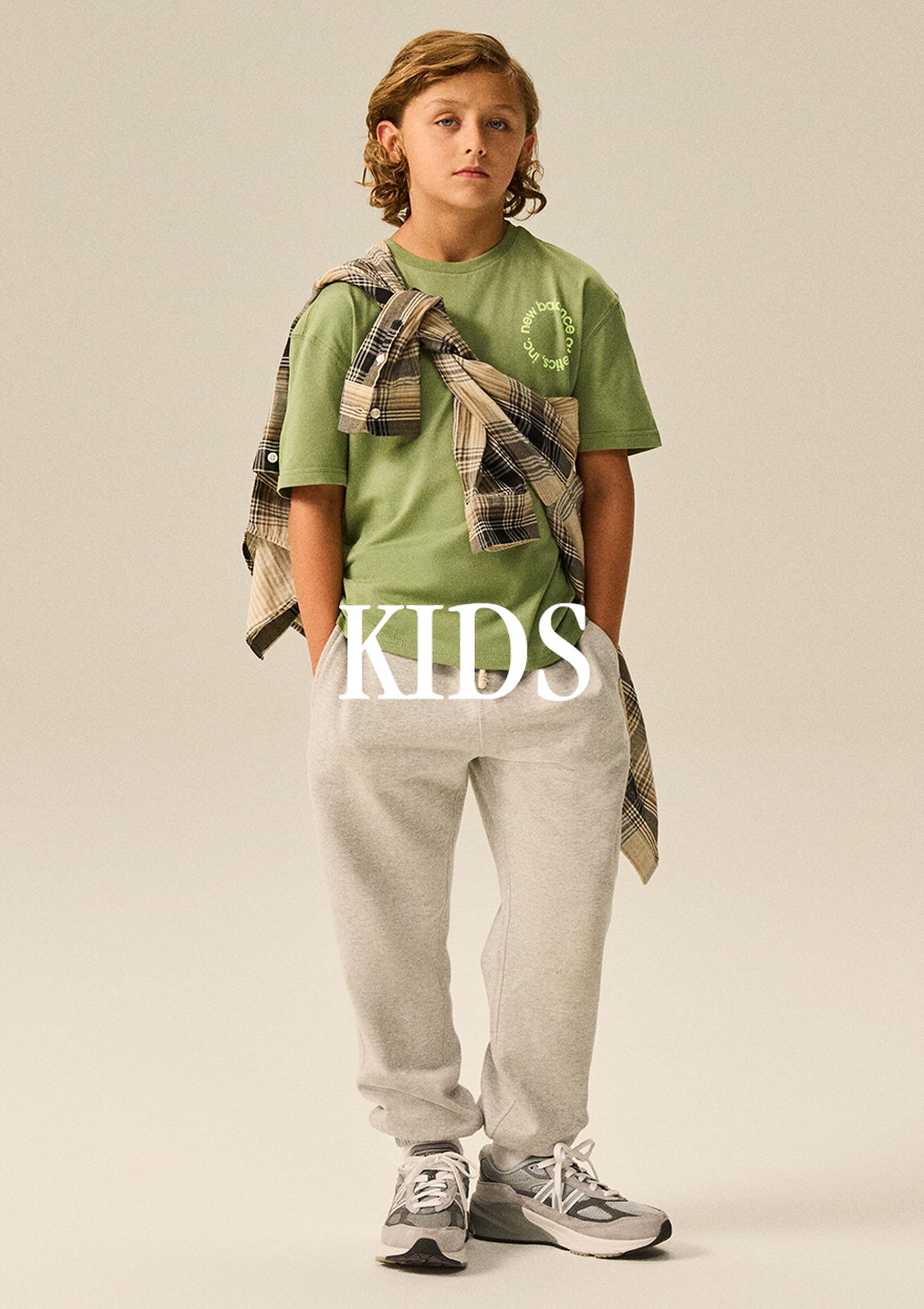 A child model in a studio fashion pose wears a green T-shirt, light gray joggers, and New Balance sneakers, with a plaid shirt tied across the shoulders and the word “KIDS” overlaid on the image.