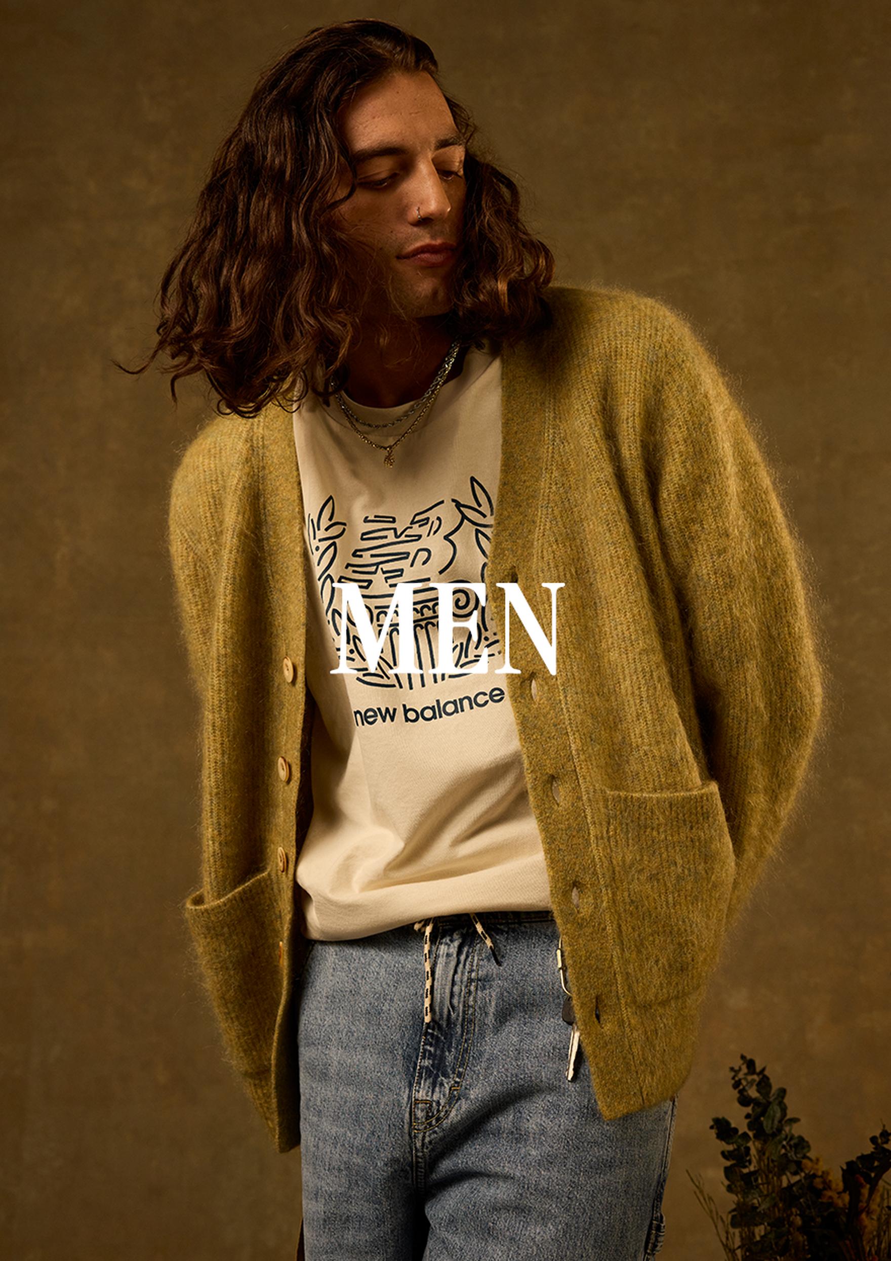 A man in a neutral-toned studio fashion portrait wears a New Balance outfit, including a black cap, an oversized cardigan, a branded T-shirt, and jeans, with “MEN” overlaid on the image.
