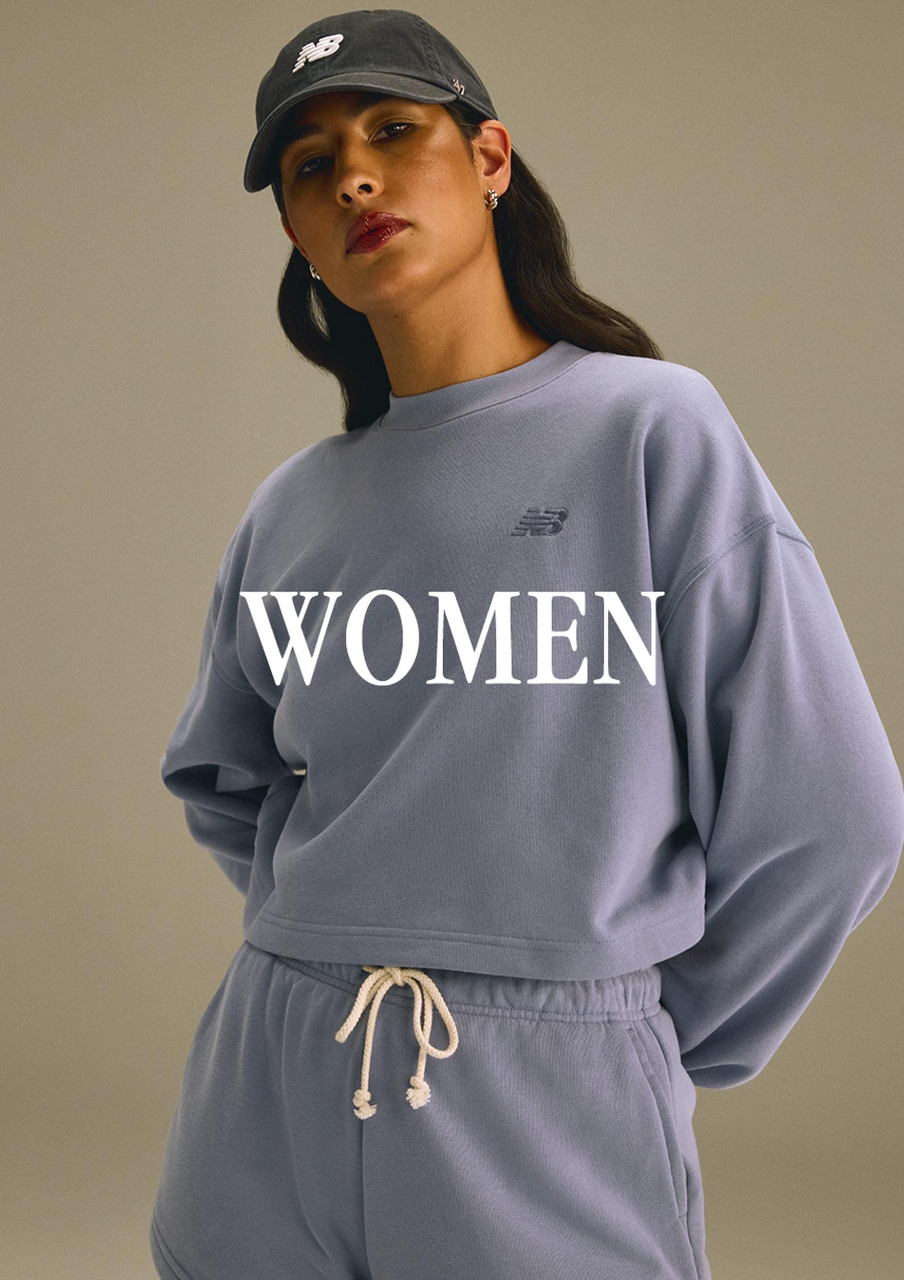 A woman wearing a black New Balance cap and matching lavender-blue New Balance sweatshirt and joggers, shown in a clean studio-style fashion image with the word “WOMEN” overlaid across the top.