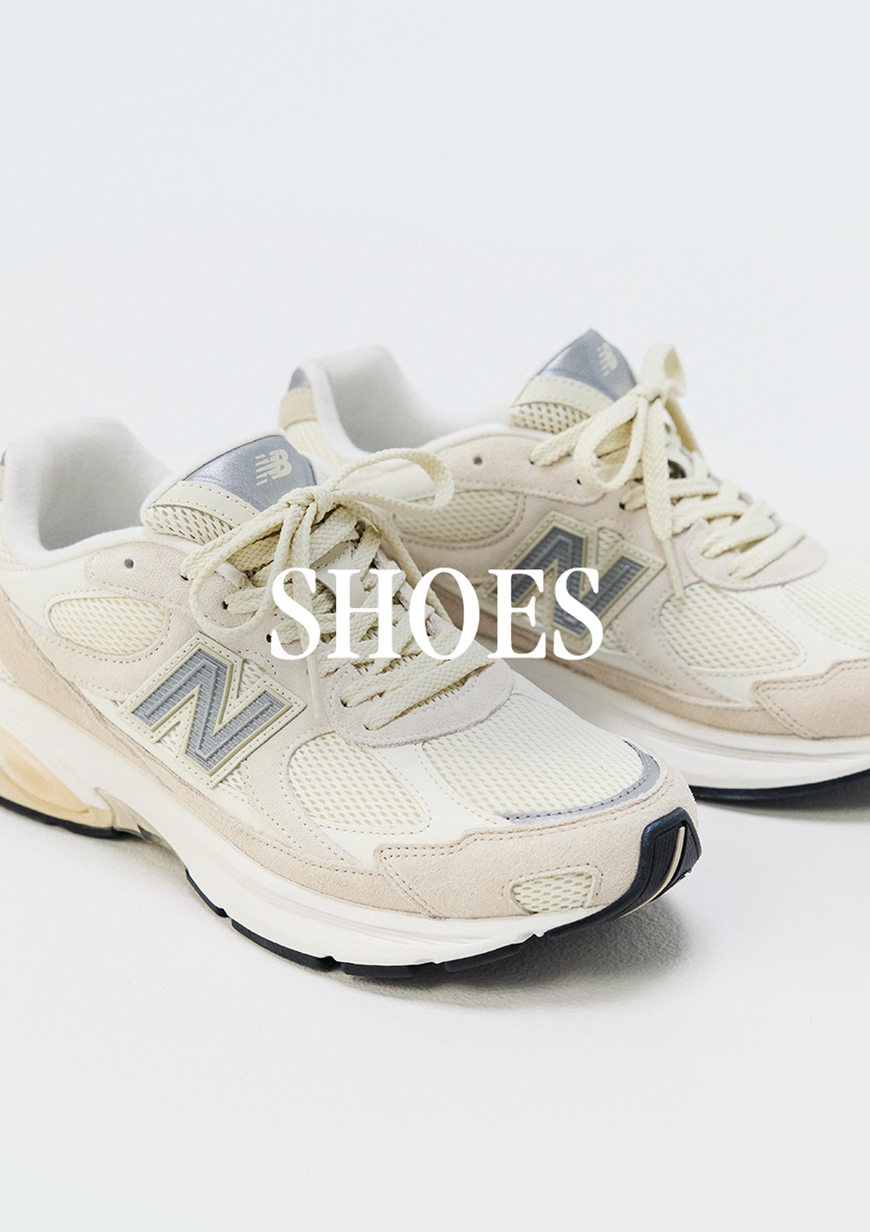 A pair of cream New Balance sneakers with blue-gray accents on a light studio background, with the word “SHOES” overlaid across the center. MENS