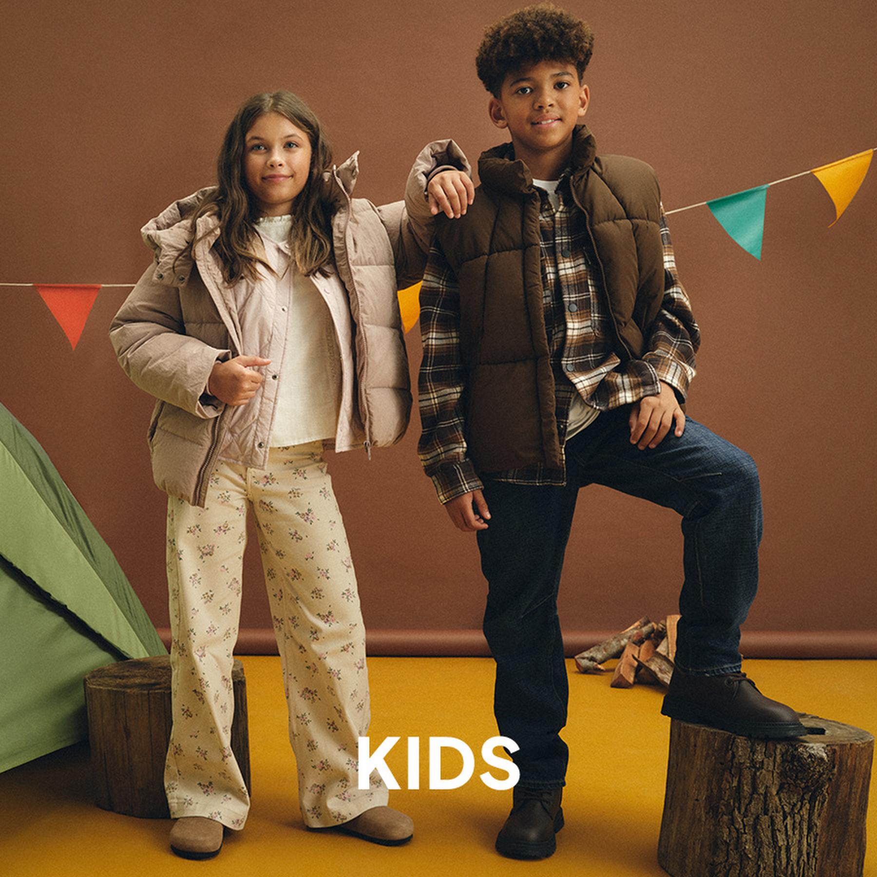 A girl in a beige puffer jacket and pink button-down skirt, and a boy in an olive green jacket and brown trousers, both wearing boots. Kids' collection.