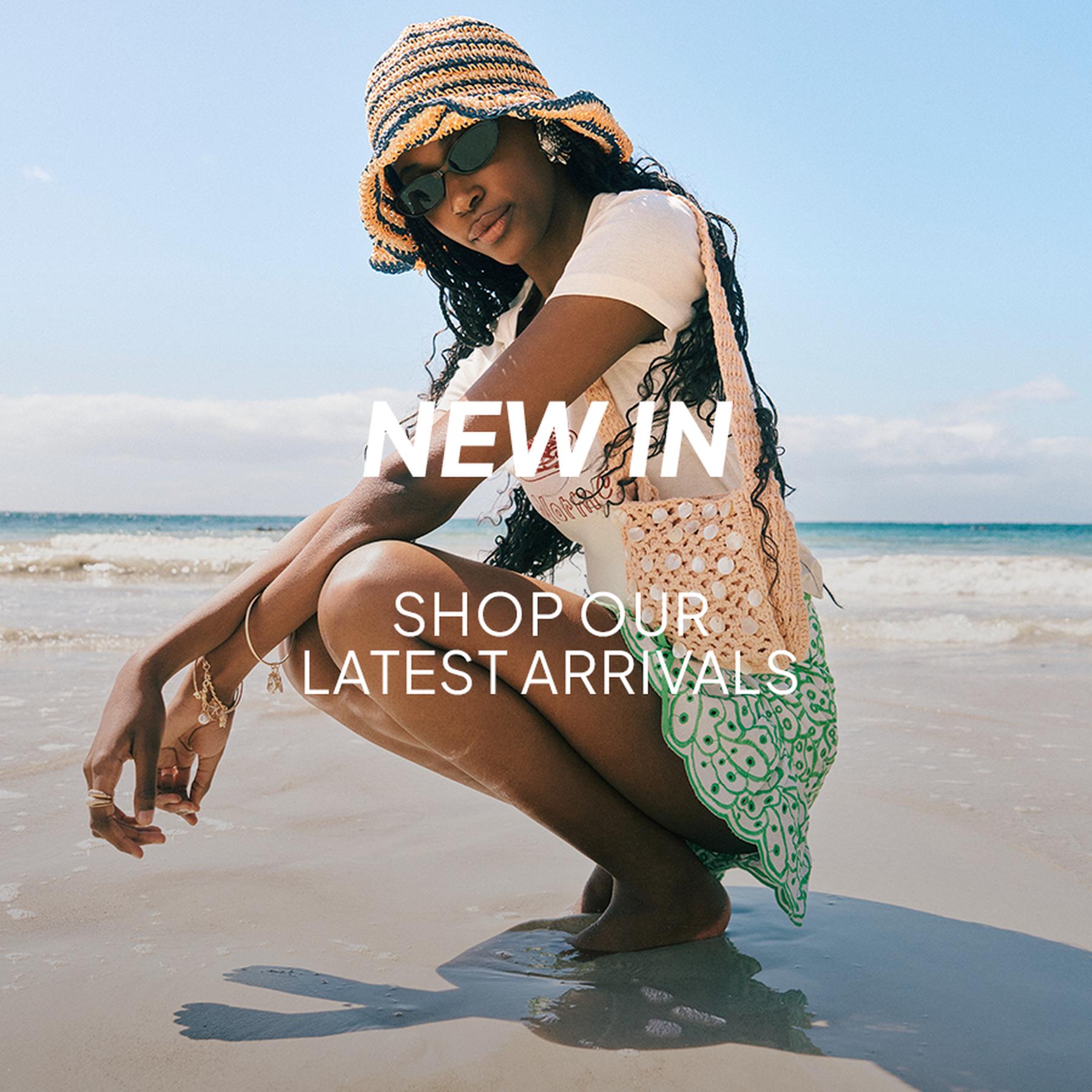 Woman standing in shallow beach water beside large rocks, wearing a light textured two-piece outfit and holding sandals, with “NEW IN” and “SHOP OUR LATEST ARRIVALS” text overlaid.