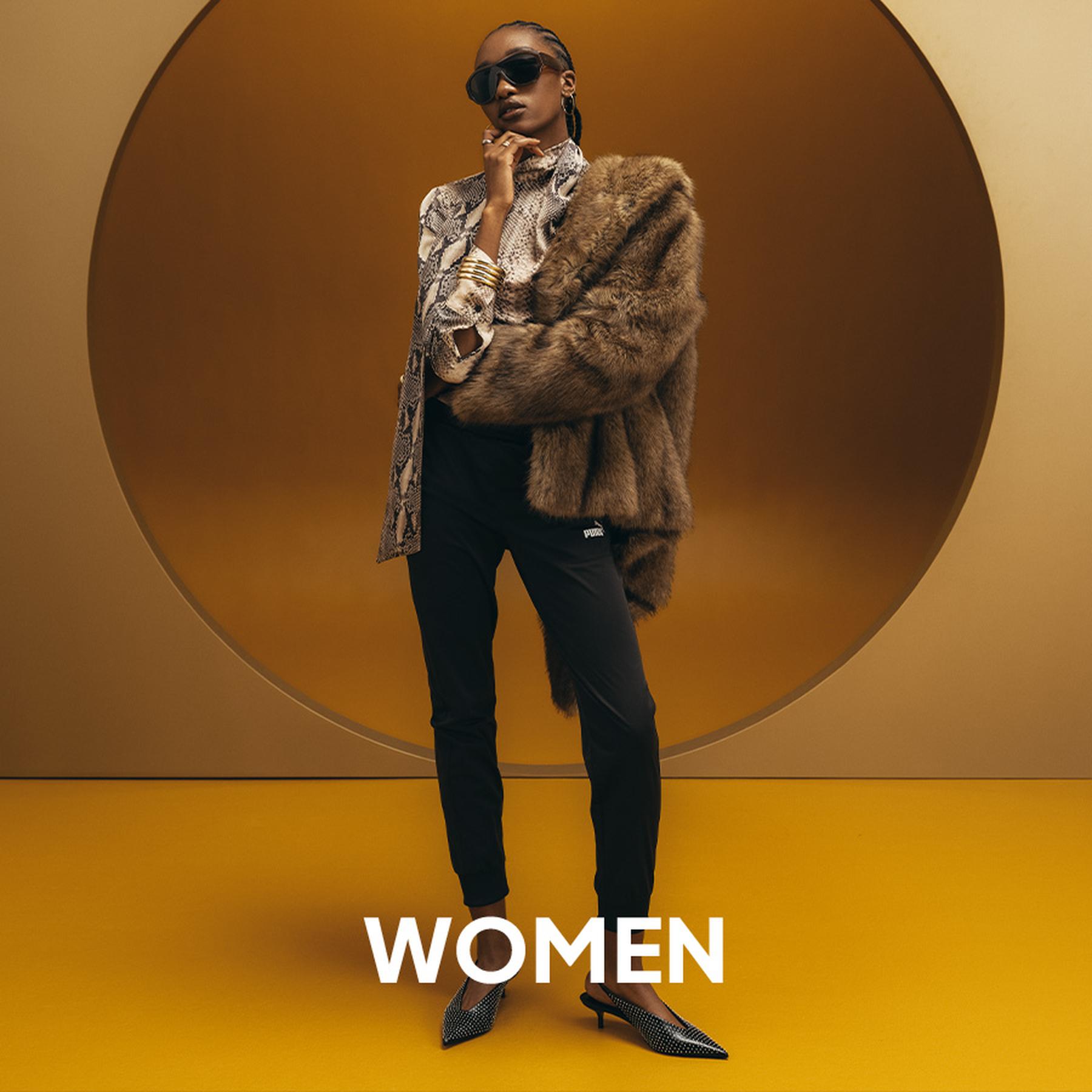 A woman in an oversized dark chocolate brown coat holding a matching suede clutch bag in a minimalist studio. Women's collection.
