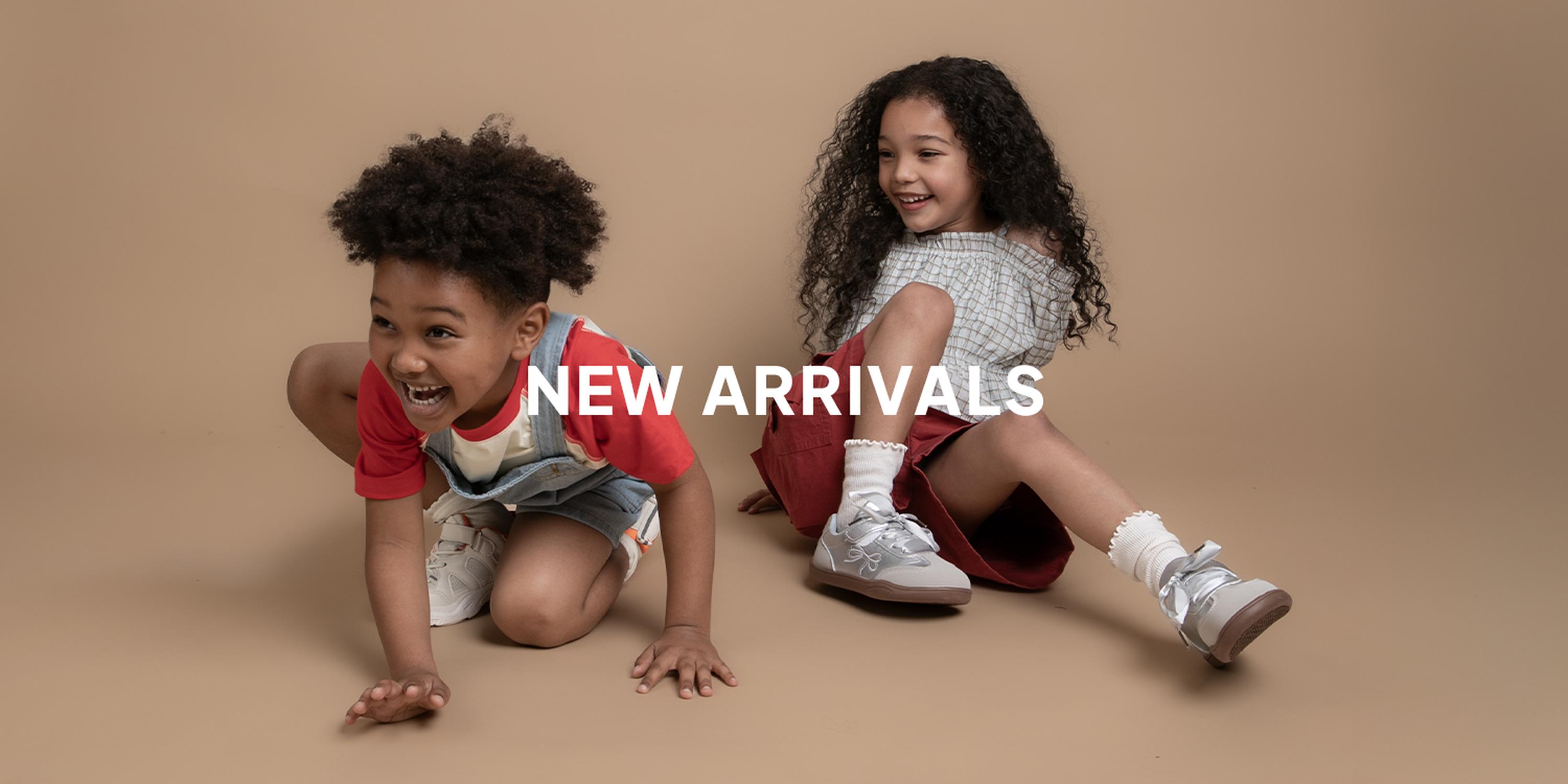 Two children posing playfully in studio against a tan background, with the white text “NEW ARRIVALS” centered over the image. KIDS CLOTHING, FASHION, ACCESSORIES, HOME