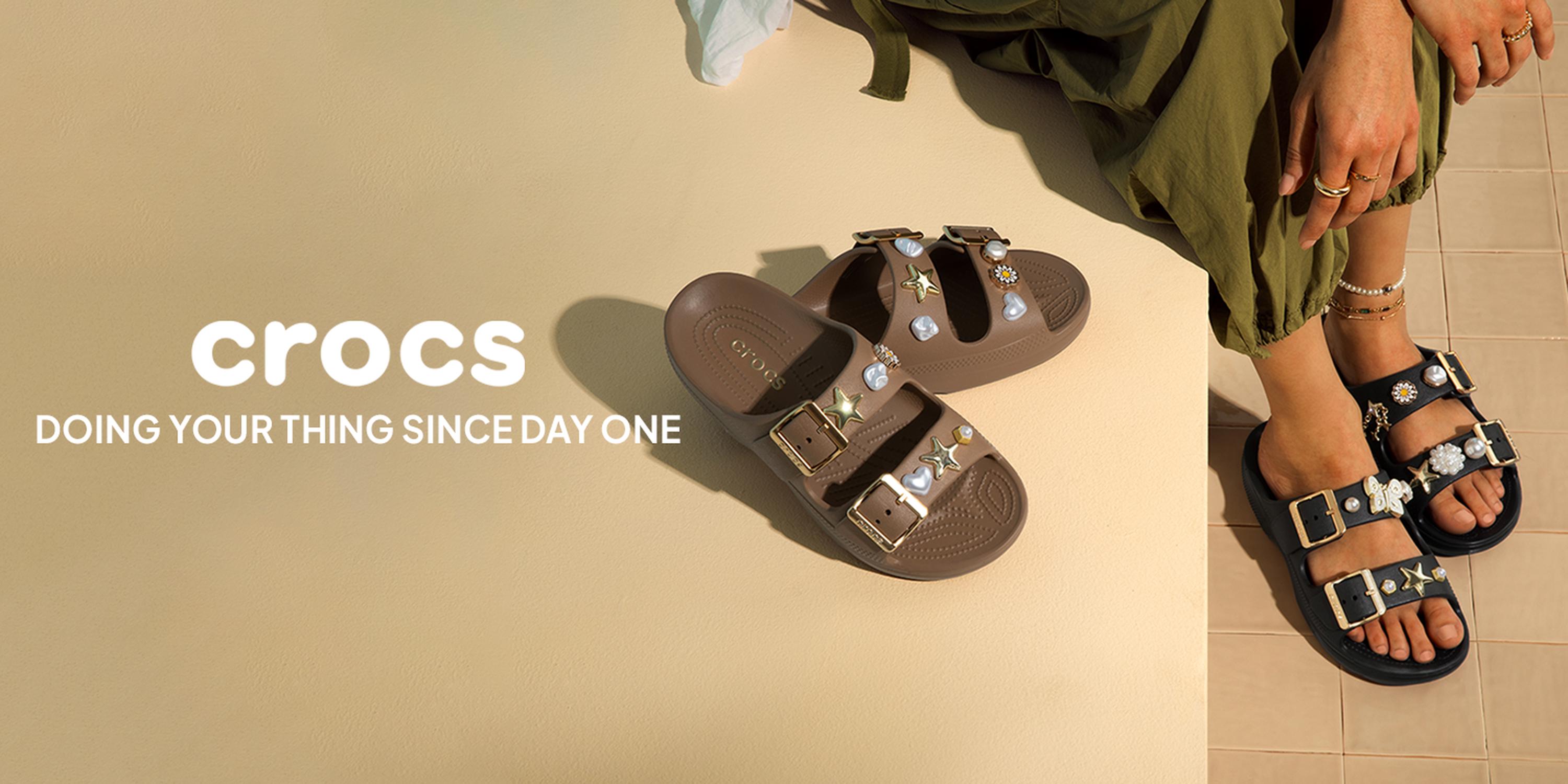 Crocs sandals ad with two embellished buckle slides in brown and black beside a model’s feet on a beige floor, featuring the Crocs logo and the slogan “DOING YOUR THING SINCE DAY ONE”