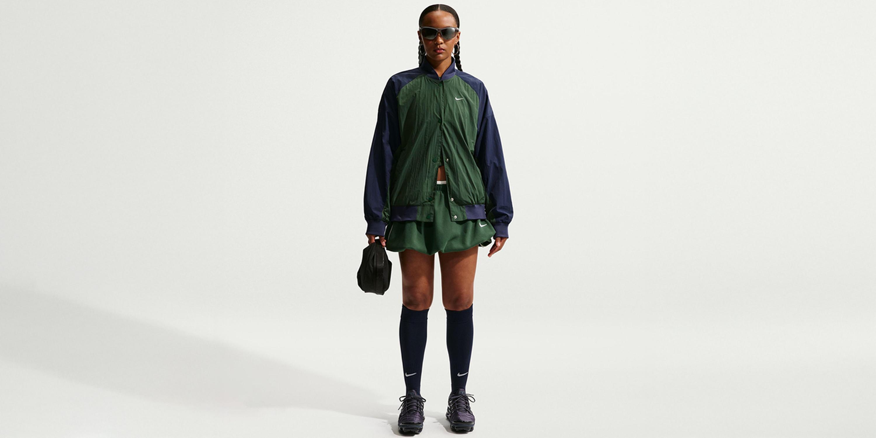 Full-length Nike fashion image of a woman in sunglasses wearing a dark green and navy Nike tracksuit with a green skirt, holding a black gym bag against a plain light grey background.