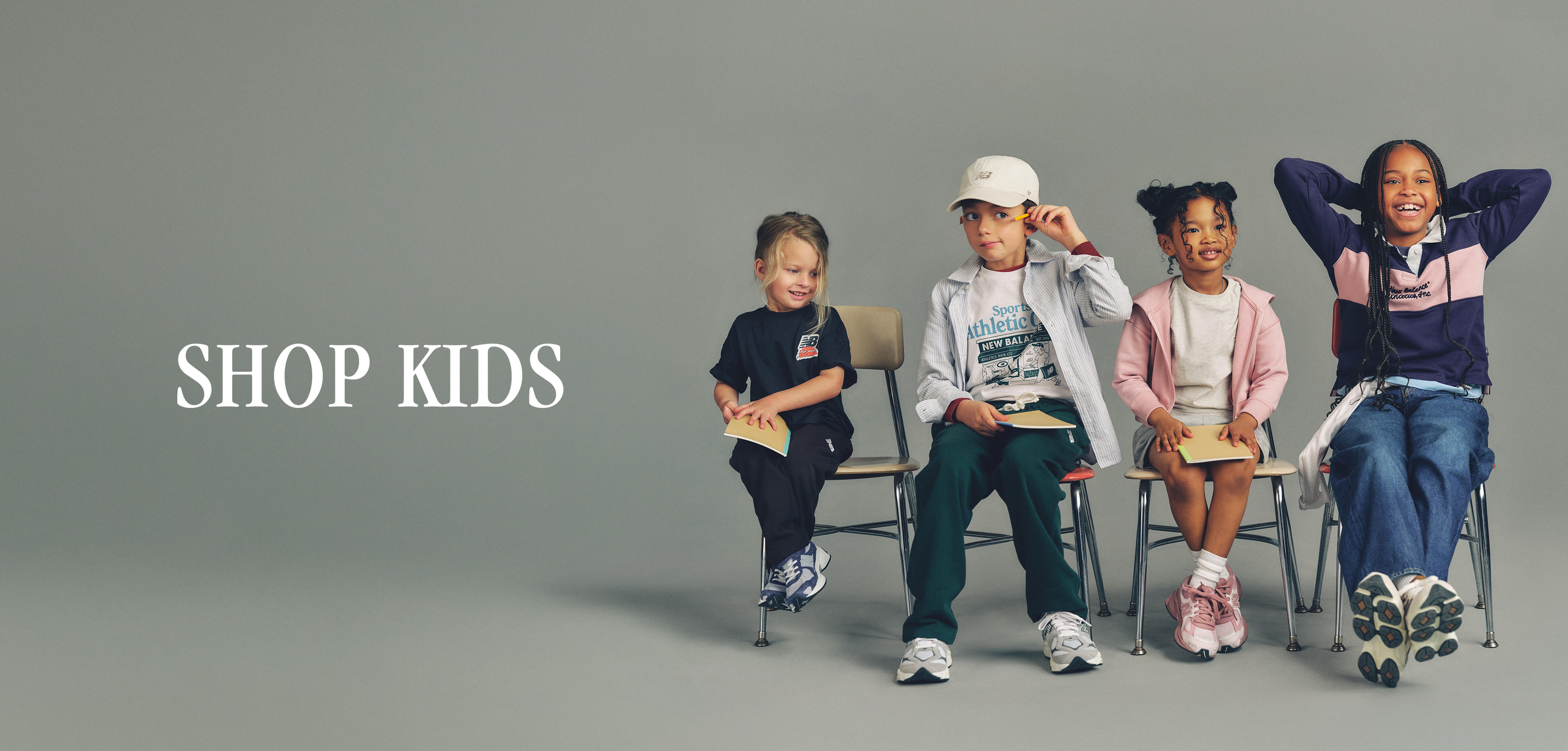 Studio-style New Balance “SHOP KIDS” banner showing four children seated on chairs against a plain gray background, wearing casual athletic outfits and sneakers; one child laughs with arms raised while the others sit relaxed.