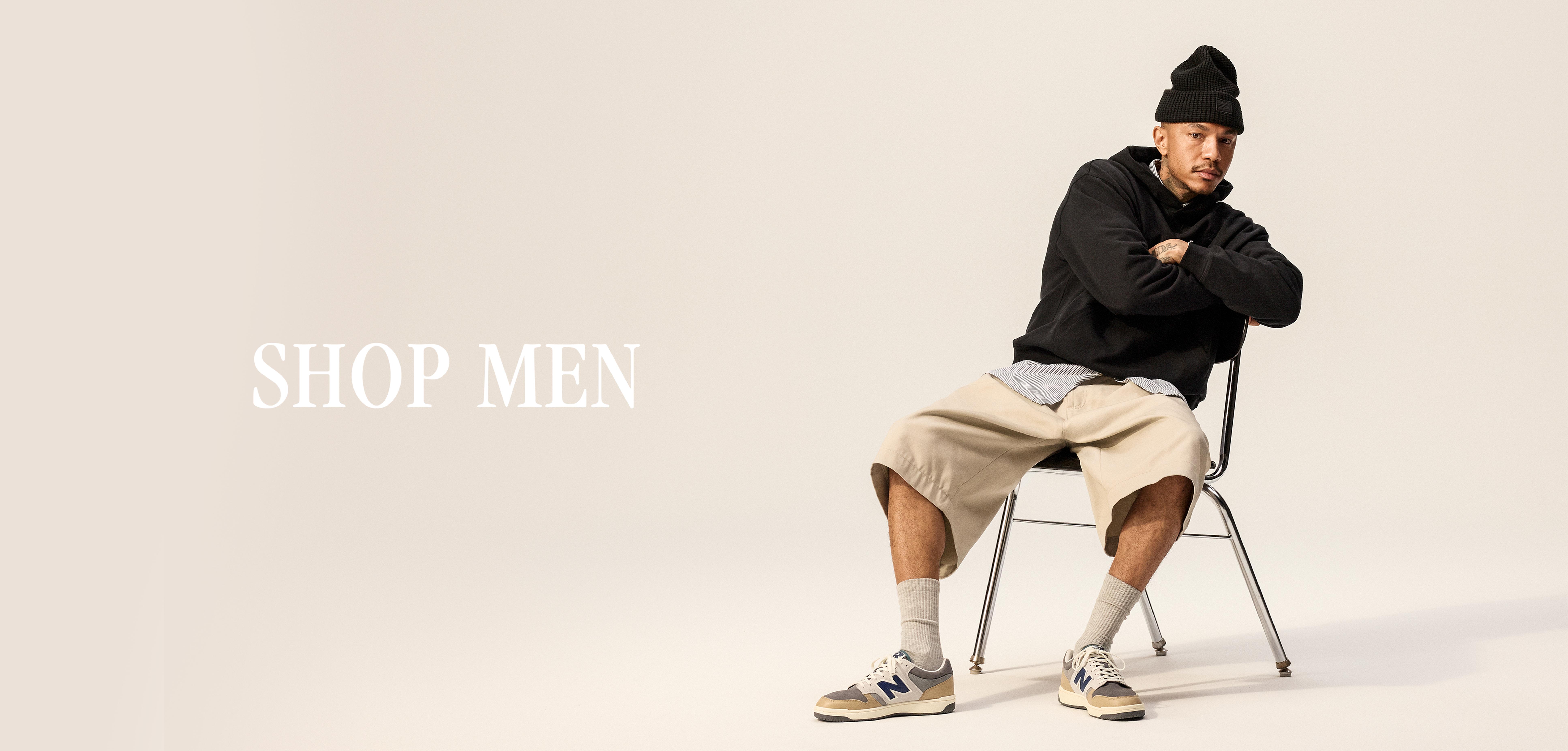 Studio fashion image of a man in a black beanie and black hoodie with beige shorts and New Balance sneakers seated on a chair, with large text reading “SHOP MEN” on a plain light background.