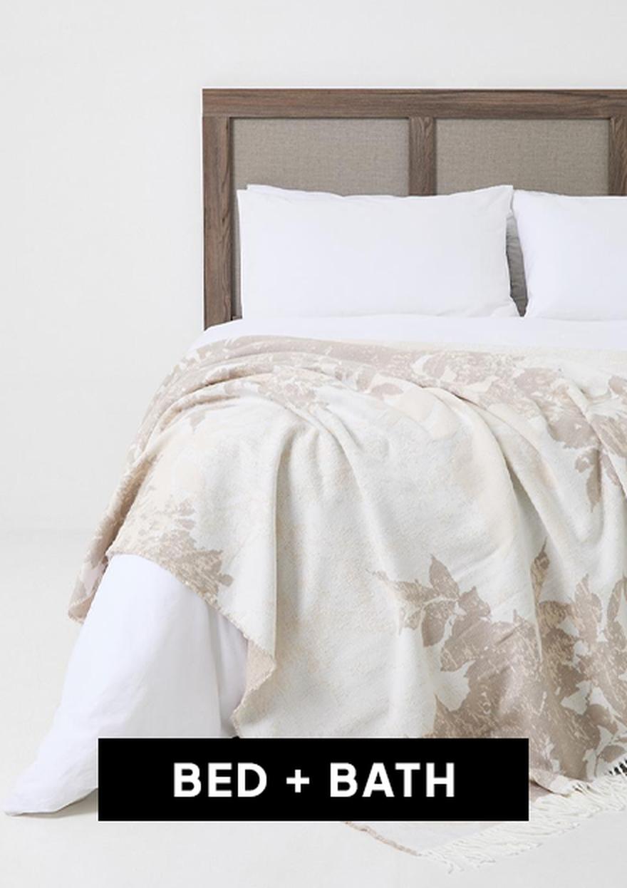 A neatly made bed with white bedding and a pink throw blanket, labeled “BED + BATH” for a Cyber Monday home and living deal.
