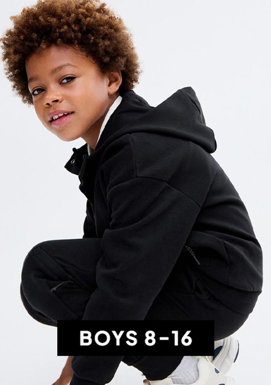 Smiling young boy with curly hair wearing a light beige T-shirt against a white background, shown as the “BOYS 8–16” Black Friday kids clothing deal.