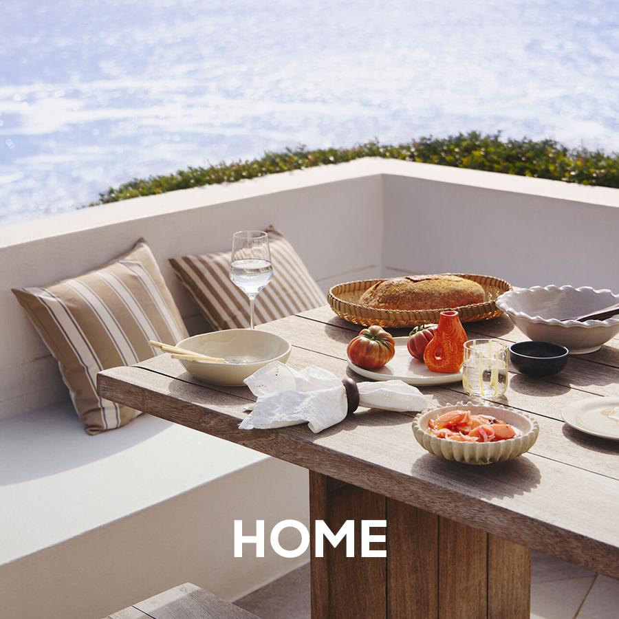 Superbalist home banner showing an outdoor dining table set on a white patio with striped cushions, bread, fruit, bowls, and glassware, overlaid with “HOME” text.