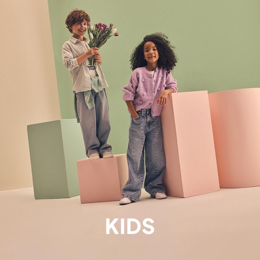 A girl in a beige puffer jacket and pink button-down skirt, and a boy in an olive green jacket and brown trousers, both wearing boots. Kids' collection.