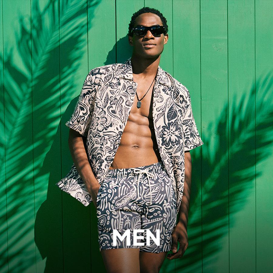 Superbalist men’s fashion banner showing a model in patterned shorts and an open short-sleeve shirt, wearing sunglasses and posing against a bright green wall, with “MEN” text overlaid.
